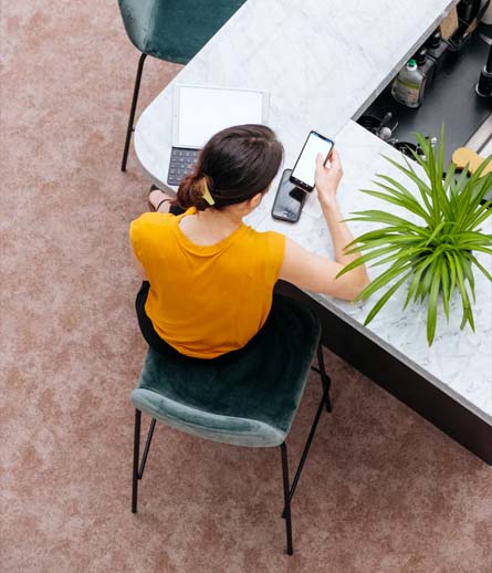 Een persoon in een geel shirt zit aan een witte tafel, geconcentreerd werkend op een laptop en tegelijkertijd een smartphone vasthoudend, wat een moderne, flexibele leeromgeving voor online Lean training en opleidingen symboliseert.